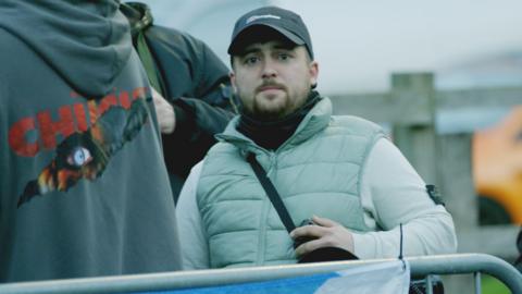 Connor Graham standing behind a metal barrier at a protest. He has a beard and is wearing a black baseball cap, light grey top and a bodywarmer.