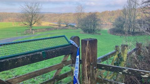 A view of the Ribble in the distance surrounded by countryside. In the foreground is a fence with police tape