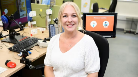 Jenny Boucher in a white top with blonde hair, sat on a black chair at a wooden desk 