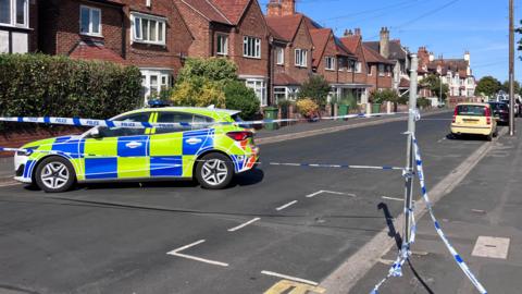 A police car is parked across a road. There are semi-detached houses in the background and blue and white police tape across the road in front of the car.
