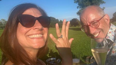 Outdoor photograph taken in a grassy park on a sunny day. Two adults sit on a picnic blanket with glasses of sparkling wine. The person in the foreground wears dark sunglasses and holds up a hand to display a silver ring on the ring finger. The second person, wearing red-framed glasses and a patterned shirt, sits opposite holding a drink. Trees and open green space are visible in the background beneath a clear blue sky.