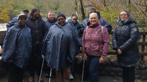 A mixed group of people stand in front of a fence by a river, posing for a picture. They all wear waterproof coats and winter clothing.