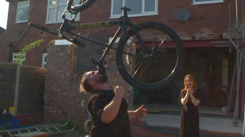 A man balances a bike on his chin outside a house as a woman laughs in the background