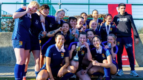 Fifteen members women's team wearing navy blue t-shirts and shorts and long socks pose on a pitch. A women at the front holds a gold cup and they all have medals around their neck, some holding them between their teeth.