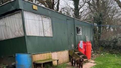 Green rusty caravan with white curtains shut. It is standing on green grass with red gass bottles and steps in front of it. Two fluffy brown dogs are stood outside the caravan.