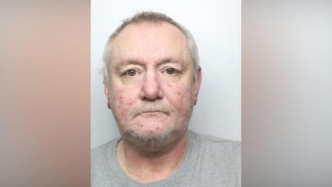 The mugshot of David Jack, a man with light grey hair and a light moustache with stubble. He is starting impassively forwards. 