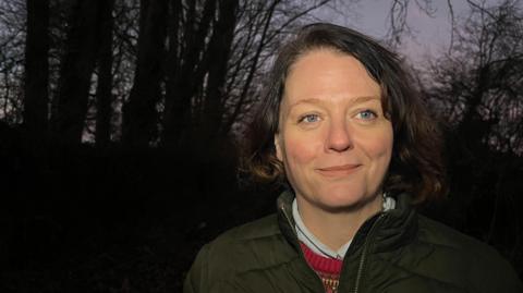 Eliza Whitaker standing in front of trees, looking past the camera. It appears to be dusk