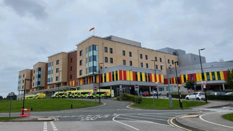 A hospital with ambulances parked outside it. The hospital building is several storeys high, and has red and yellow cladding on the lower part.