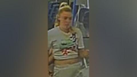 CCTV footage of a blonde woman wearing a grey crop top and joggers. Her hair is tied back in a ponytail and there are train seats visible in the background. 