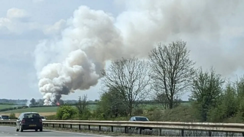 A big plume of smoke rises from a building in fields next to the M4, with cars driving past it, with trees to the right side of the motorway