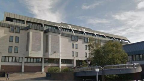 Maidstone Crown Court is a large building with windows visible on five floors. A pedestrian walkway can be seen just in front of the building, along with some trees and shrubs.