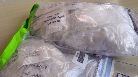 A close-up view shows several transparent plastic evidence bags resting on a light-coloured surface. Each bag contains white powder and is sealed, with handwritten notes and codes visible through the plastic. One bag features a bright green sealing strip, while another has a barcode label. In the background, a partially visible official label or document is attached to one of the bags, reinforcing the fact these bags are catalogued samples.