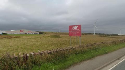 The New Balance Athletic Shoe factory. The picture is taken from the A596. The factory is on the left. A red sign naming the company is in the middle near the road. Two existing wind turbines stand on the right.