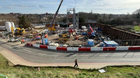 An elevated view of the £58m Energy Centre under construction on a sunny day. The site is next to a road and a range of heavy vehicles are building the frame of the structure.