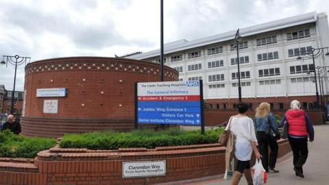 Leeds General Infirmary hospital entrance. In the foreground, there is a curved red-brick wall with a blue NHS sign listing directions to hospital departments, including Accident & Emergency, Jubilee Wing, Visitors Car Park, and Dental Institute. Below the wall, a smaller sign reads “Clarendon Way.” Several people are walking on the pavement toward the hospital building.