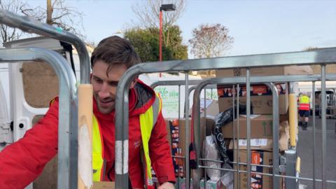 A man with brown hair in a red postal jacket and a high-viz vest reaching for a package. He is behind a trolley full of parcels, next to a white postal van.