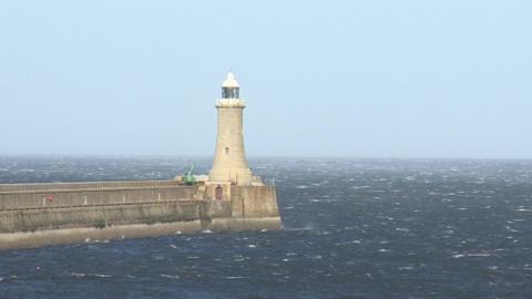 Tynemouth's North Pier. The lighthouse structure at the end of the pier is made of beige bricks, with white and glass sections at the top. There is a green crane parked in front of it. The pier is made of grey bricks and is surrounded by the choppy waters of the North Sea.