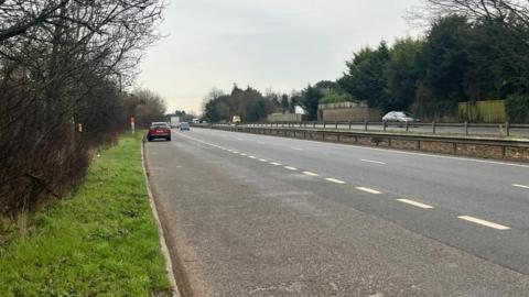 A picture of a lay-by at the side of a dual carriageway There is grass to the left of the lay-by. The sky is grey, and there is a red car parked in the lay-by. 