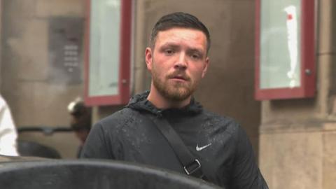 Jordan McMillan is wearing a black jacket. He has a brown beard and brown hair.