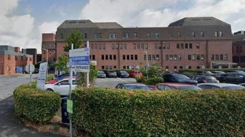 View of Scunthorpe General Hospital building with cars parked in front of it and a hedge in the foreground. The hospital is made from red brick.