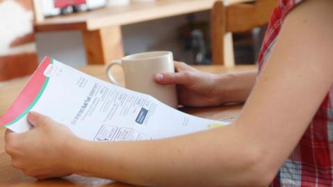 A woman wearing a checked top sits at a wooden table, holding a bill in one hand and a mug in the other.