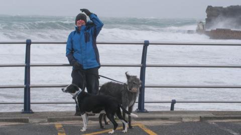 A man wearing a blue all-weather rain jacket is standing in front of a stormy sea in Cornwall. He is in front of railings and is looking at the camera and has two dogs on leads 