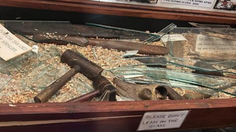 A broken cabinet with old artefacts exposed and a sign which says please don't lean on the glass