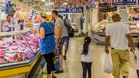 People in a market hall indoors. Several people are in between market stalls including a butchers with meat on display. A woman and a man look at the meat while a man and a girl walk away from the camera, the man carrying a bag in his left hand.