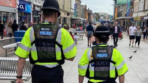 A stock photo of a male and female police office on patrol in Southend. Their backs are facing the centre and they wear yellow high visibility police jackets under a black mesh police vest. Both officers also wear black policing hats.