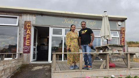 A man and woman standing on wooden decking next to a wooden bench with a white parasol. In the background is a cafe with a wall of windows and and open glass door. A light green sign with white writing reads "Kate's Cafe"