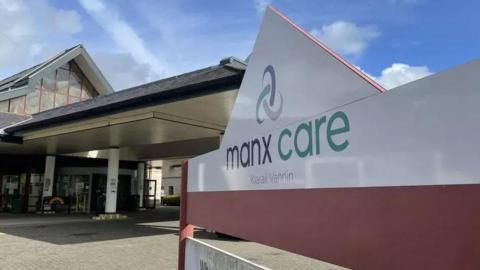 A Manx Care sign in front of the entrance of Noble's Hospital. The sign has blue, green and grey writing and the Manx Care logo. The entrance has revolving doors with a long canopy held up by pillars.