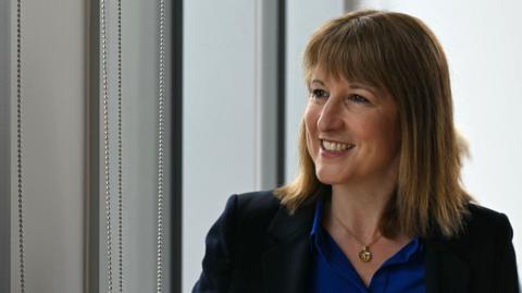 Rachel Reeves looks to the side smiling - she has shoulder-length brown hair and is wearing a black blazer with a blue shirt and a gold necklace