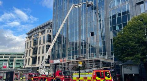 A hydraulic aerial platform is by a tall building. Four fire engines are in front of the building on the image.