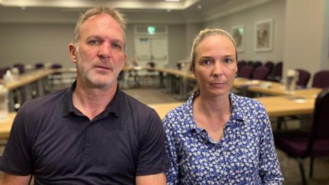 Sarah Hawkins, who is wearing a flowery top, next to her husband Jack who is wearing a polo shirt, sat in a large room with a conference style table behind