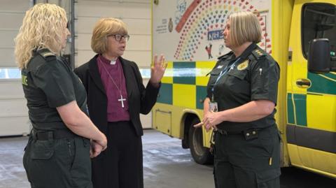 Archbishop of Canterbury Dame Sarah Mullally (centre) wearing dog collar and cross speaking to two Secamb female staff next to an ambulance