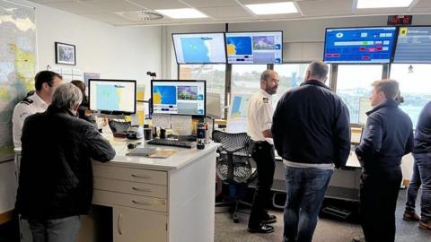 A view of the control centre at Ports of Jersey for coastguard operations. Several members of the public are seen with coastguard officers who are wearing white shirts, which have black and gold shoulder epaulettes. There are a number of screens showing maps of Jersey.