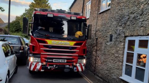 A fire engine squeezes between a stone wall and a series of cars parked on the other side of the road. Trees and a pole in the background.