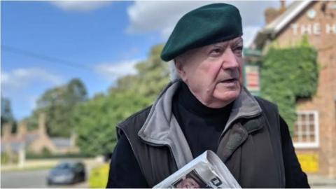 Tony Martin wearing a beret and coat, clutching a folder and a newspaper. He is standing outside a property covered in ivy