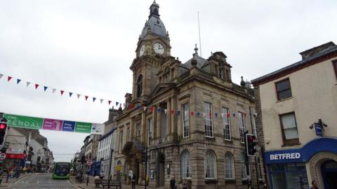 Kendal Town Hall. An ornate sandy coloured stone building with an arched entrance and pillars each side of a first floor balcony. It has a domed clock tower, with bunting stretching across a busy street running alongside.