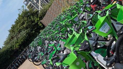 Rows of bright green Lime rental e‑bikes lined up diagonally. These bikes extend into the distance beside trees and a fence.