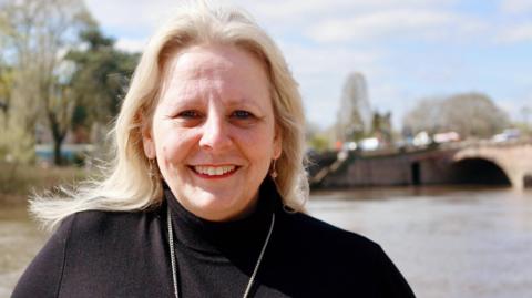 Elena Round wears a black turtle necked jumper with a silver chain. She has long wavy blonde hair.