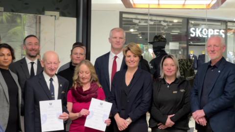 Two rows of people standing smiling for the photo - with Carns in the middle on the back row. A man and woman on the front row are holding certificates. 