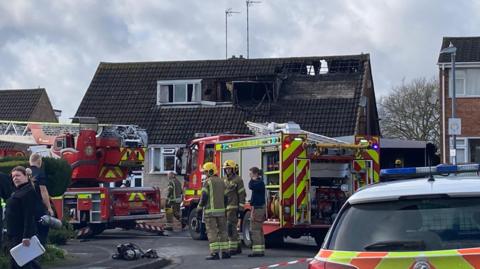 A semi-detached two-storey house with severe fire damage with firefighters and fire engines in front next to police cars