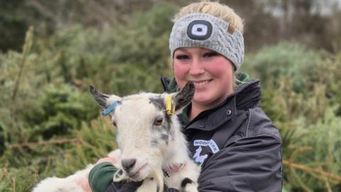 A woman wearing a grey headband and a black jacket with Noddy's Ark written on it - holding a white goat, smiling for camera.