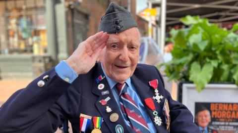 An older man, in a military cap, with numerous badges and medals on his blazer, salutes at the camera. There is a poster behind him featuring his name and a picture of him