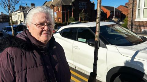 Maria Teresa Rodriguez has her grey hair tied back and wears glasses and a purple duffle coat with a black-fur lined hood, standing outside her white car which is parked on double yellow lines outside her home on Wellington Street