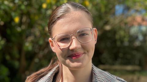 Madi Faulkner-Hatt stands outside on a sunny day and smiles directly at the camera. She has dark hair that has been tied back behind her head, she wears glasses and a chequered jacket. 