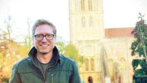 A man in a green jacket and glasses smiles at the camera, there is a church building in the background