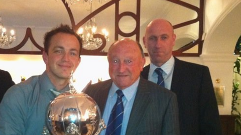 Brian Keen, an older man, is pictured centre wearing a grey suit and a blue tie. He is holding a trophy. To his right is his son, Tony, a tall man with a shaved head, and to Brian's left is James Phillips from 鶹ҳ Radio Cumbria. He has dark hair. They are all smiling.