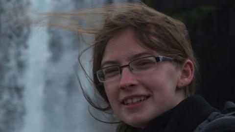 Headshot picture of a woman wearing glasses and brown hair who is smiling. Her hair is tied back but is windswept. She is stood outside and is wearing a black coat.